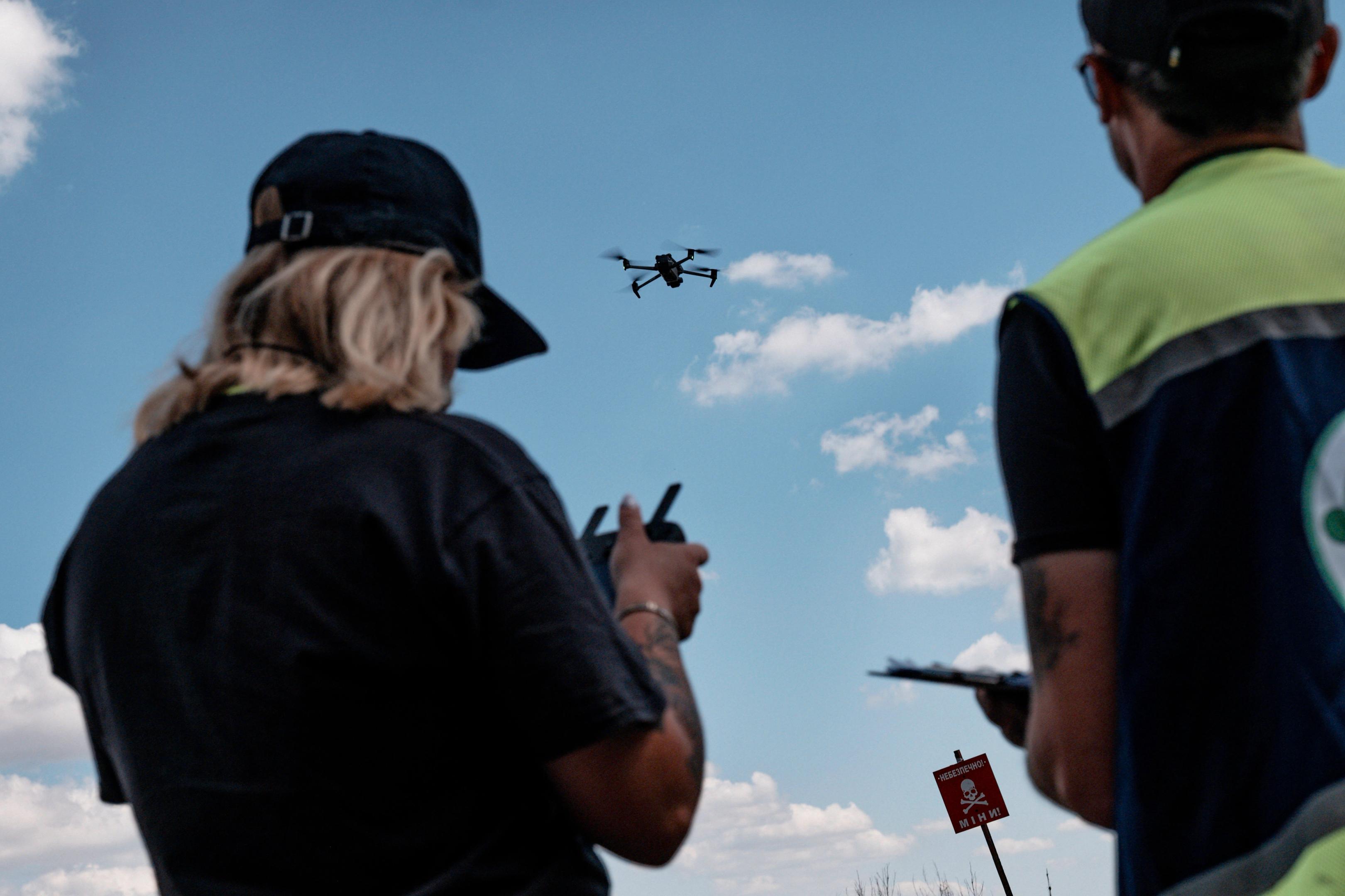 Two people standing outdoors control a flying drone, watching it hover against a blue sky with scattered clouds. One person holds a remote controller while the other appears to observe or take notes.