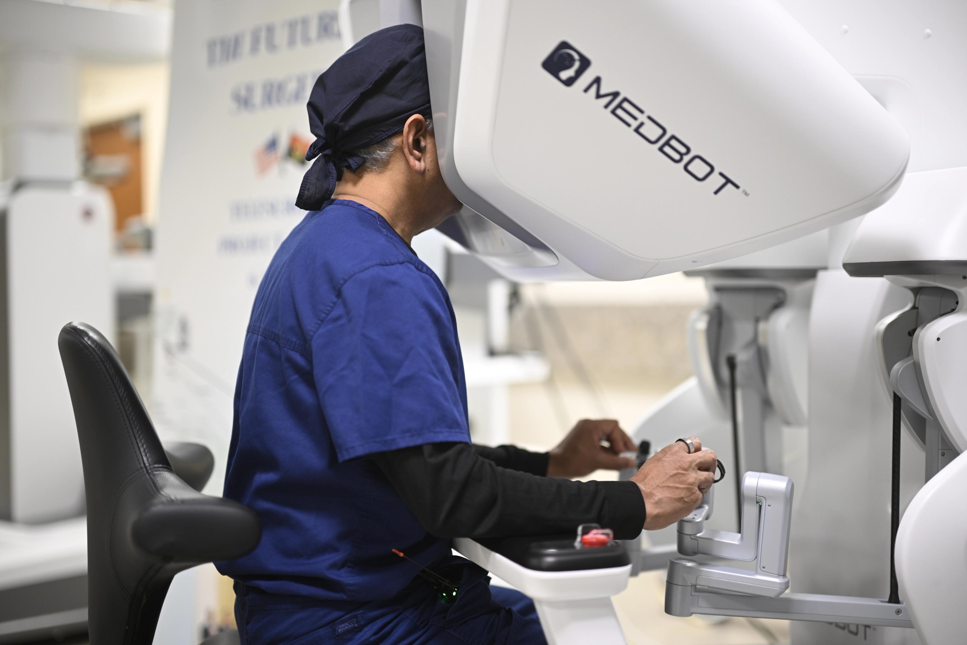 A person wearing blue surgical scrubs sits at a telesurgery console, operating a robotic surgical system labeled “MEDBOT.” The individual is using handheld controls while looking into the console, demonstrating remote surgical operation technology.