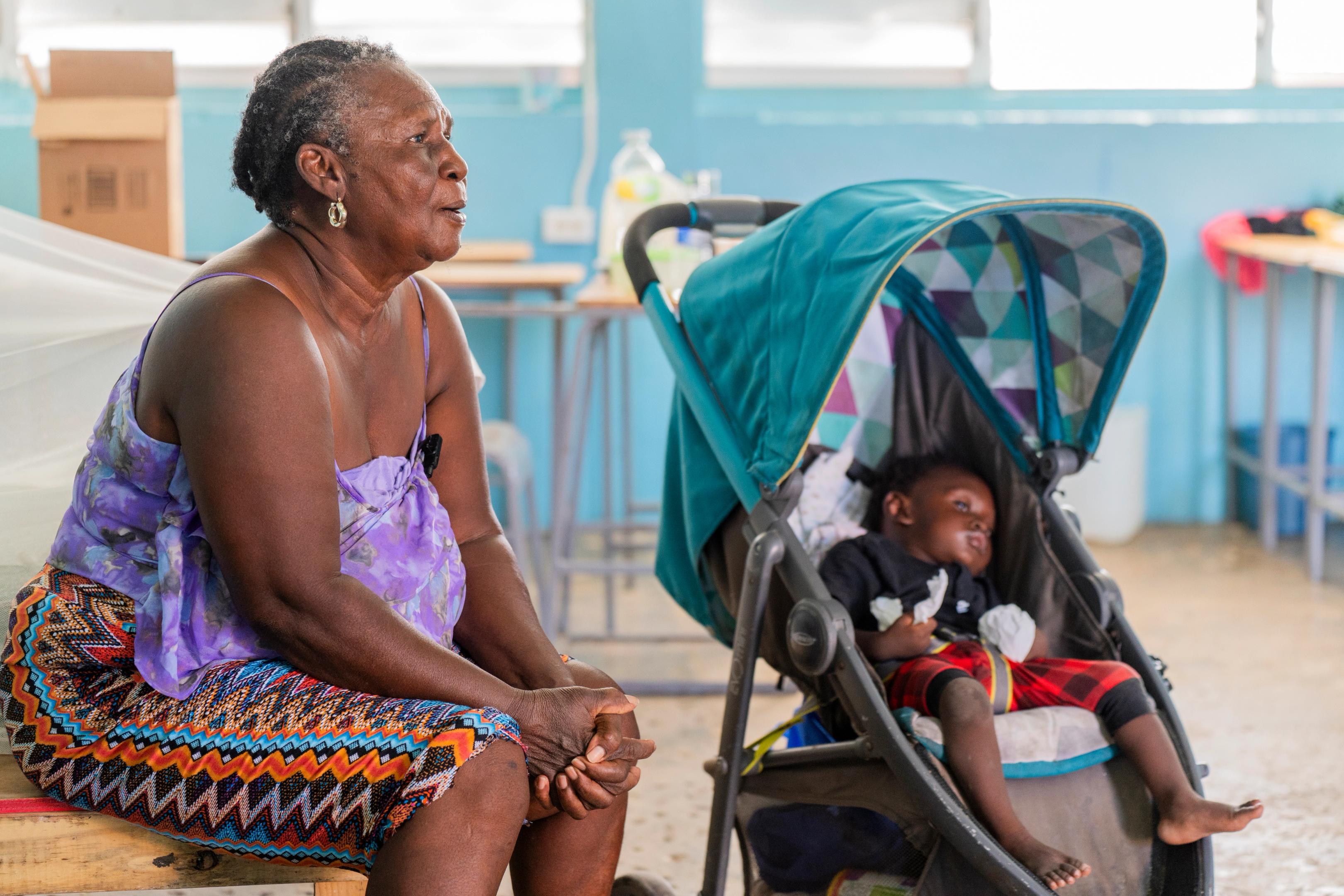A woman sits on a bench in an indoor shelter space beside a baby in a stroller. The room has bright blue walls, tables and supplies in the background, and natural light coming through high windows.