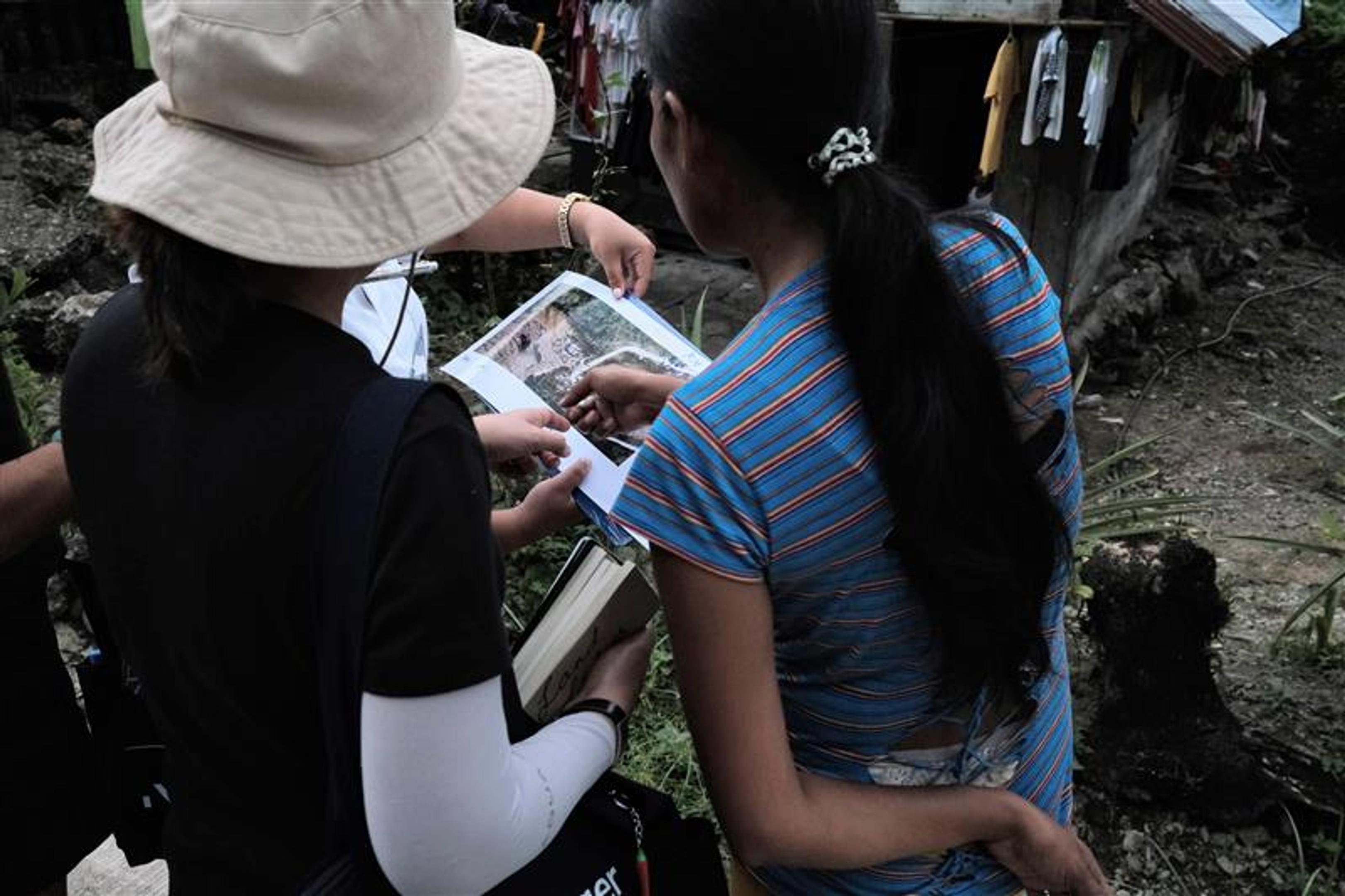 Three people stand outdoors near a small house in a rural area, looking down at a printed map or aerial photo that one of them is pointing to. The person in the foreground has long hair in a ponytail and is wearing a blue striped shirt. Another person wears a hat and carries a shoulder bag. Clothes hang to dry in the background, and vegetation surrounds the area.