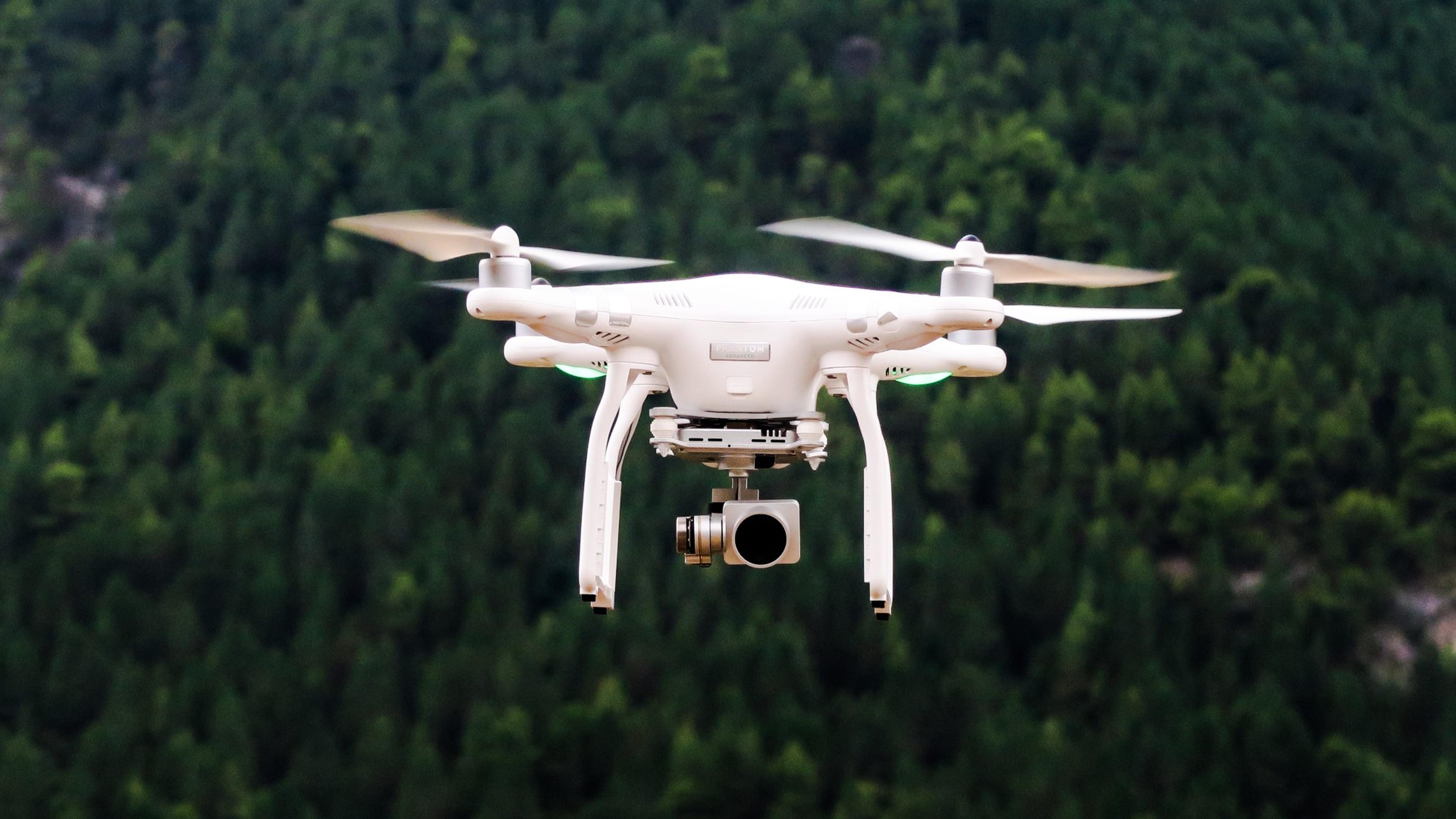 Hvit drone som flyr i lufta mot en skogsbakgrunn