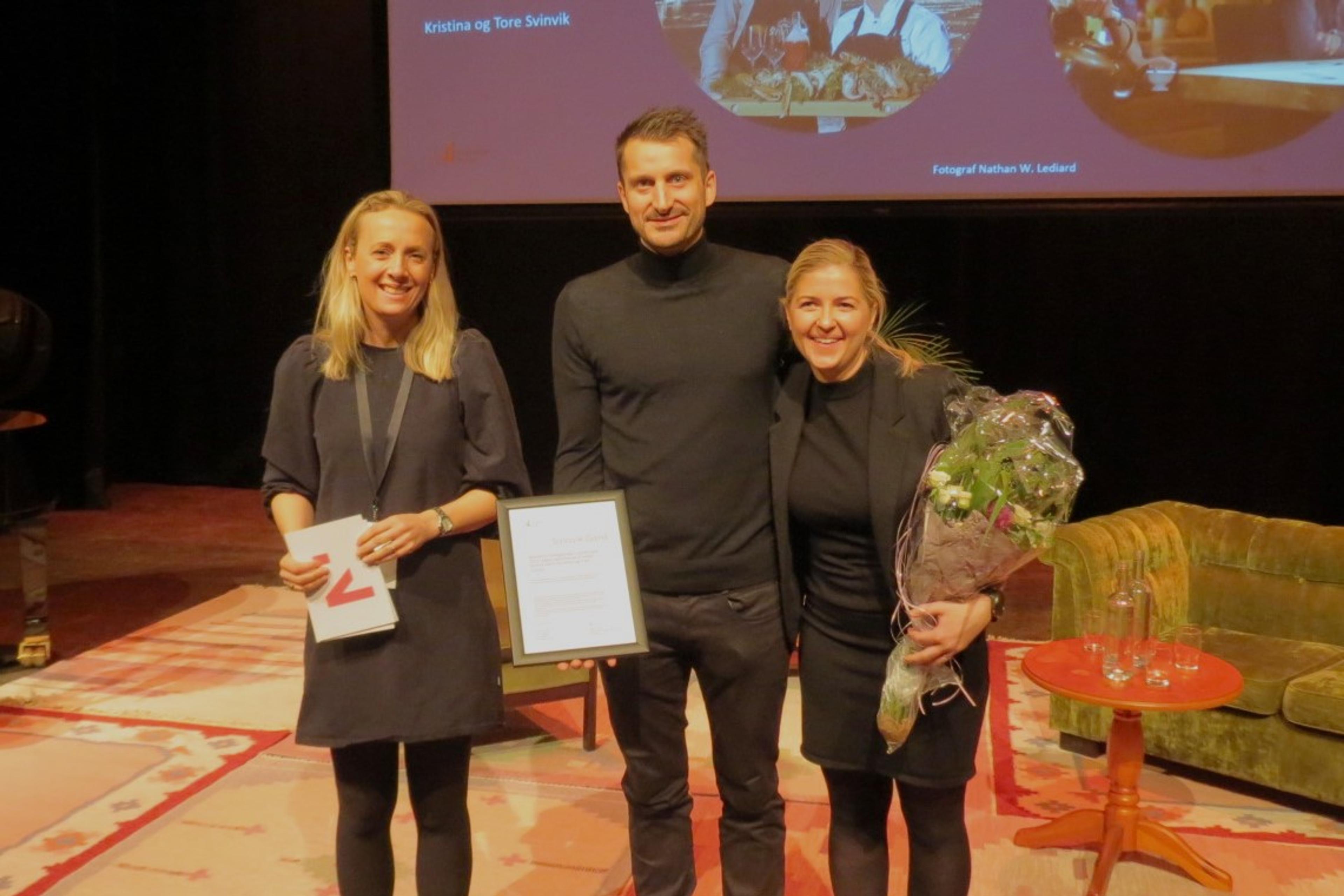 Ei kvinne og en mann mottar diplom og blomster for BU-prisen av ei kvinne med talekort fra Innovasjon Norge i hendene