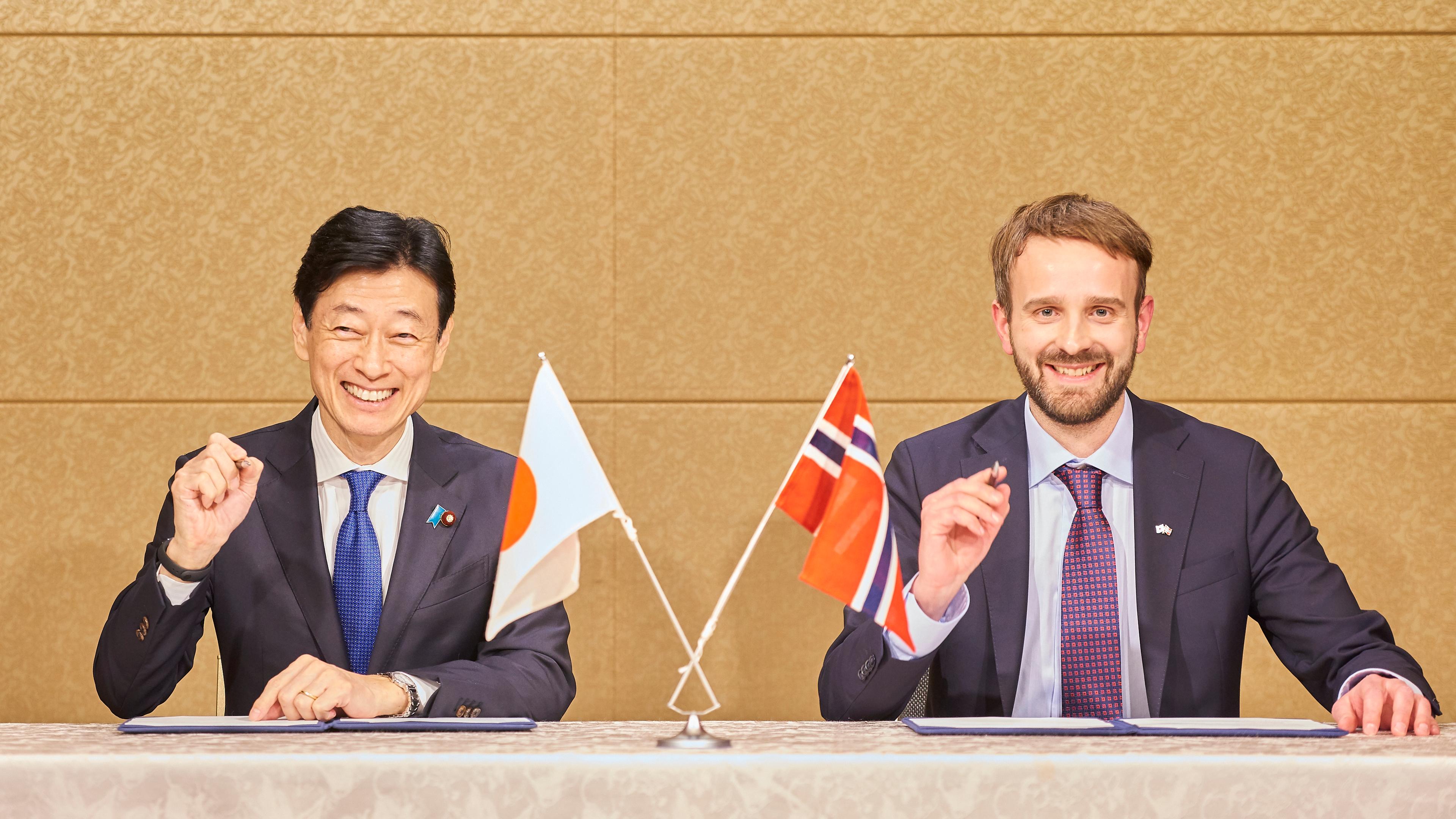 Japans handelsminister og Vestre holder opp hver sin penn mens de smiler før de skal signere