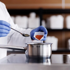 En person i hvit frakk og plasthansker heller rødt pulver nedi en kasserolle