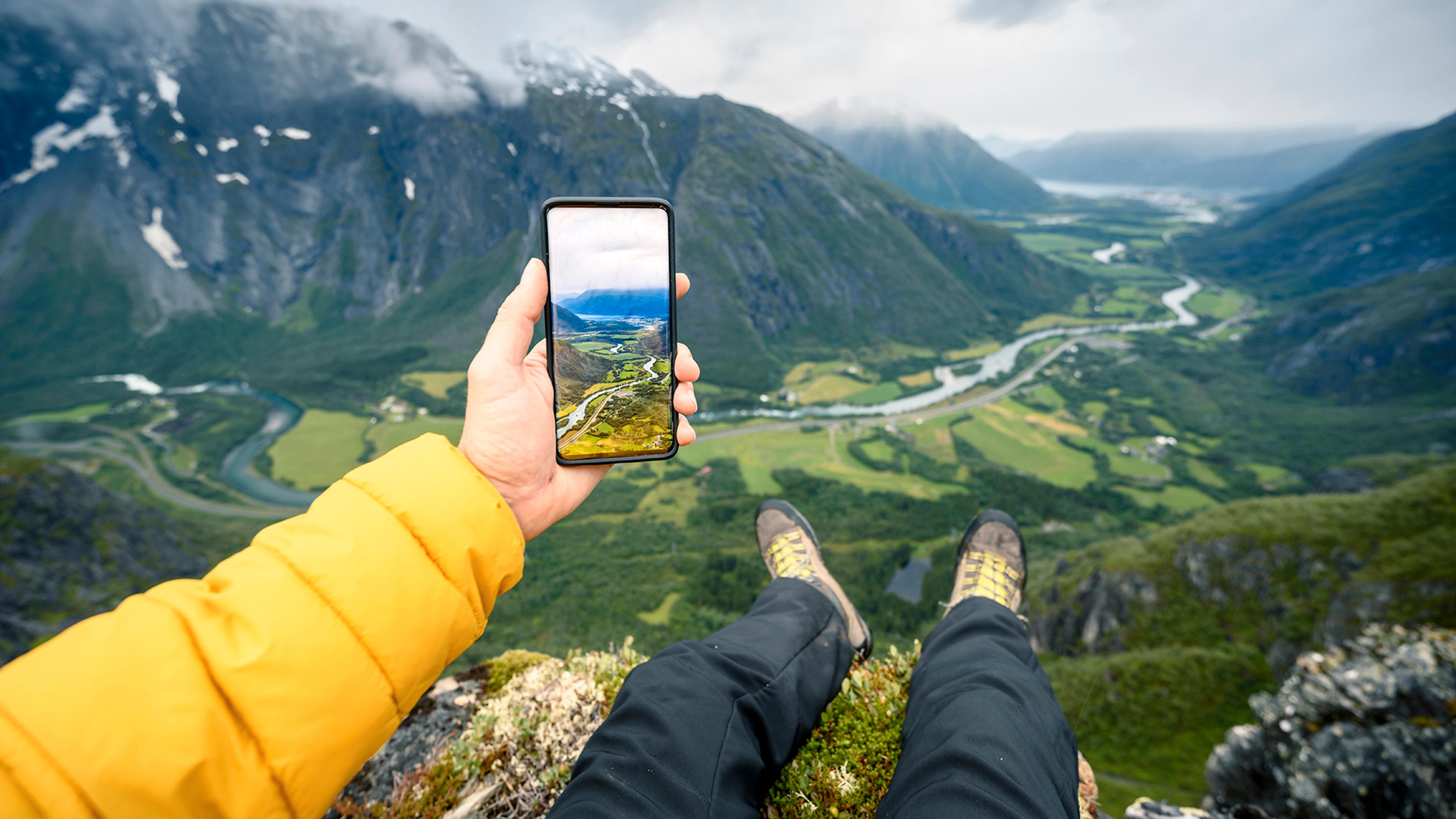 Person som sitter på fjelltopp og ser på bilde hen har tatt av utsikten med mobilen