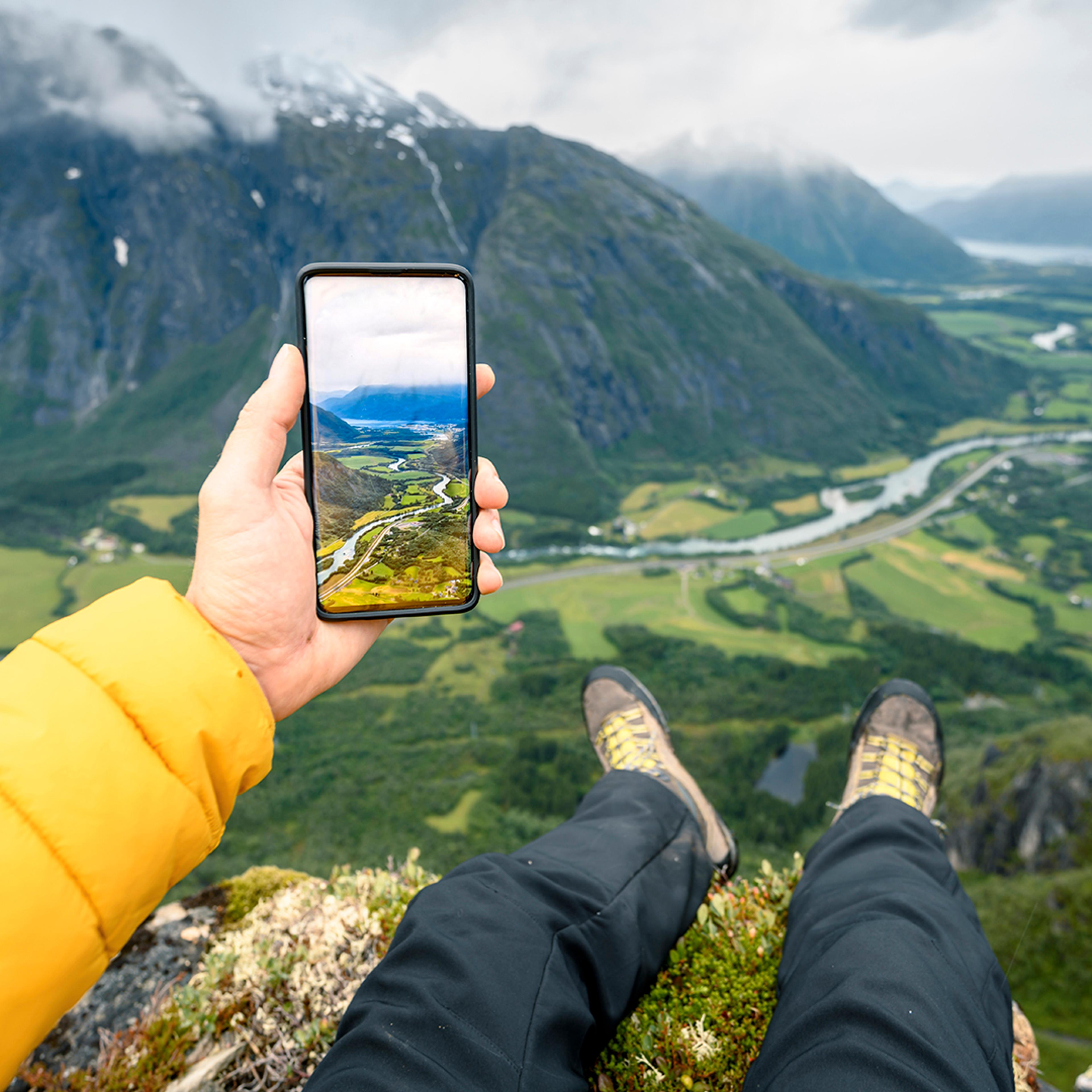 Person som sitter på fjelltopp og ser på bilde hen har tatt av utsikten med mobilen