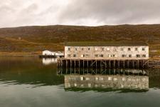 Bilde av gammel bygning som står på påler i vannet. Blankt hav, grå himmel.