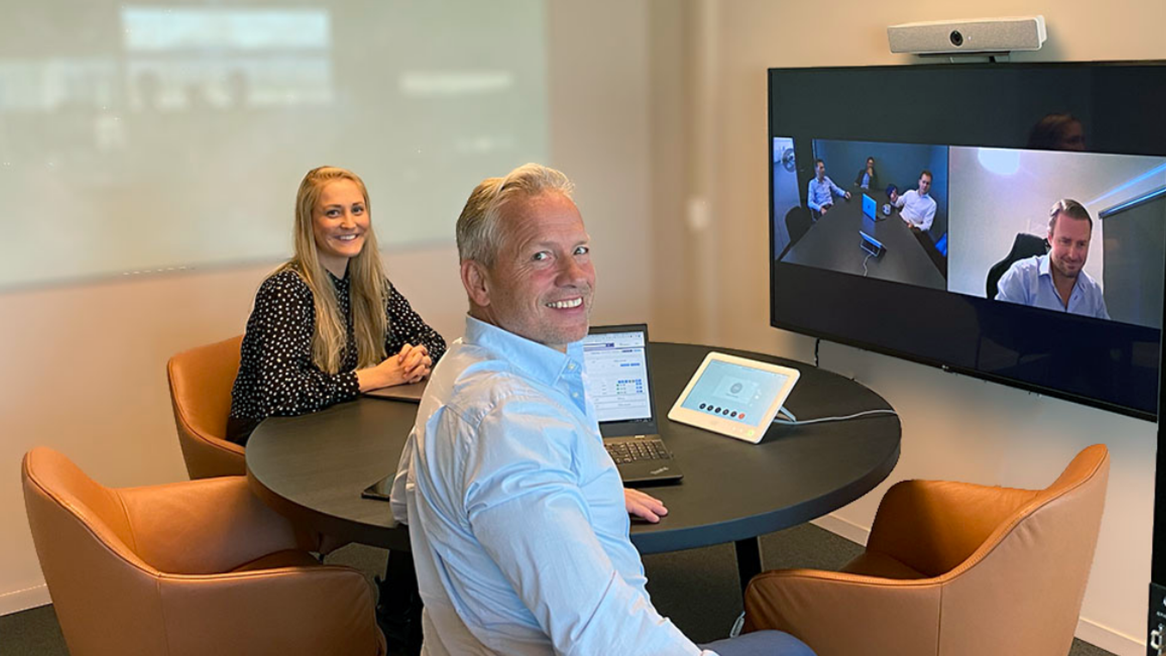 To mennesker sitter sammen i et rom og deltar i et digitalt møte fra en skjerm på veggen
