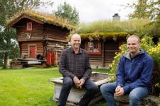 To menn sitter sammen på et tun med et stabbur i bakgrunn