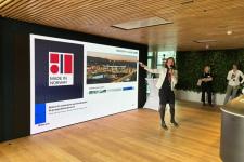 A woman in a white blazer and a microphone in front of a giant screen