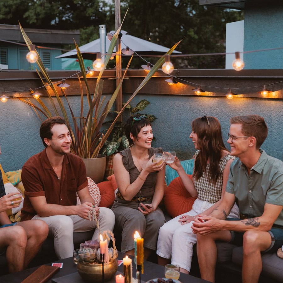 Cozy backyard patio at night strung with warm glow string lights, where friends lounge on pillows and blankets watching a movie on a projector screen, surrounded by plants, candles, and rattan chairs.