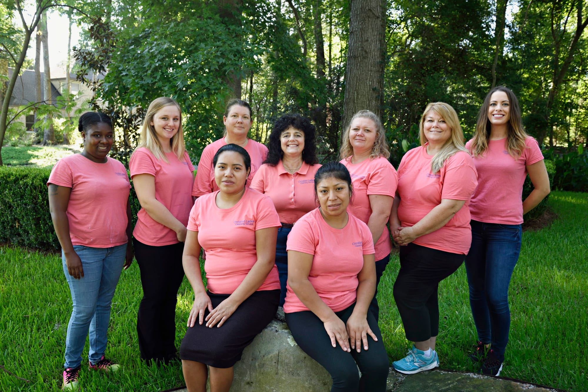 Nine women in pink company shirts posing together on a lawn in front of trees.