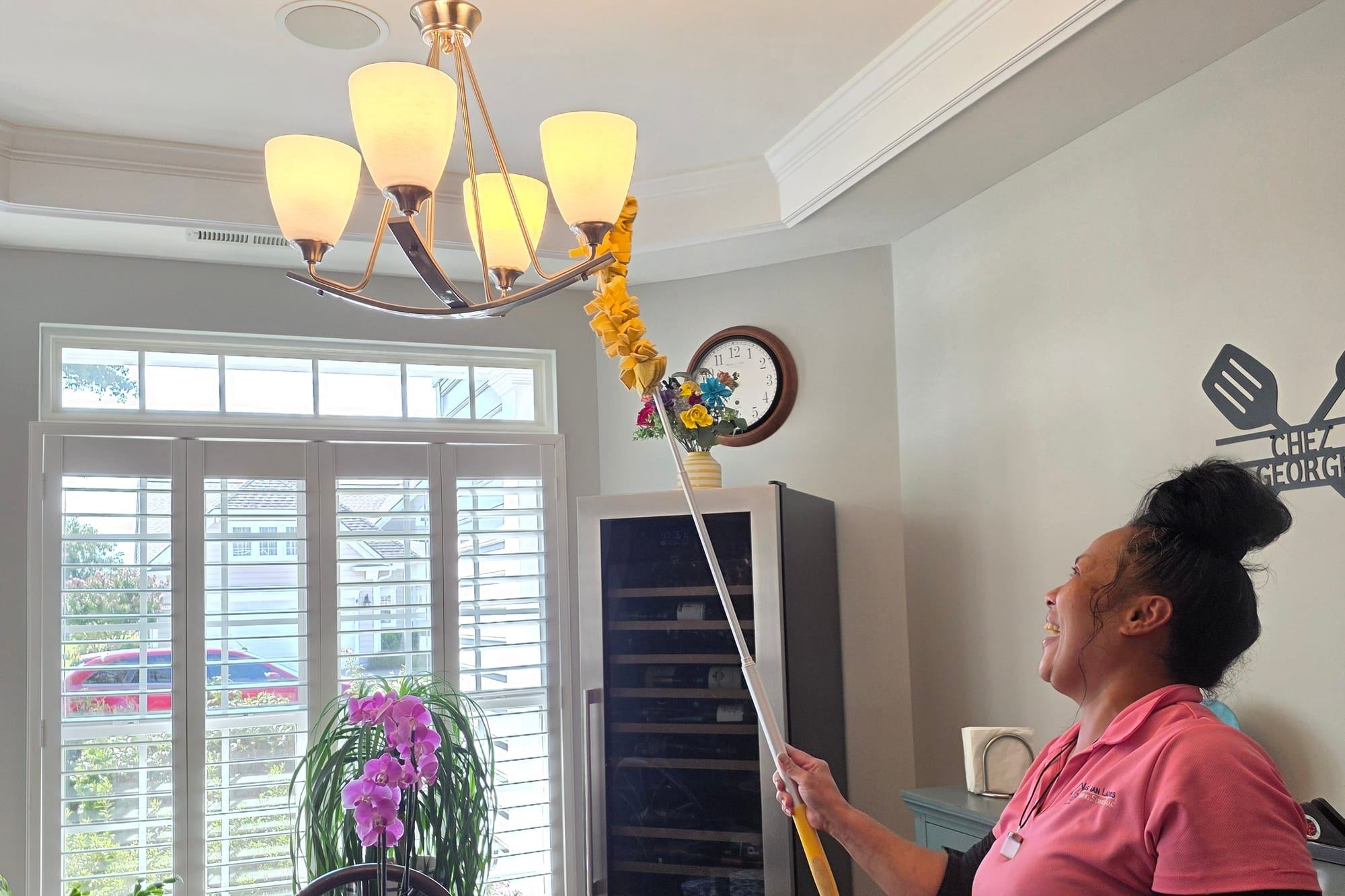 A smiling cleaner in a pink polo dusting a dining room chandelier with an extended duster.