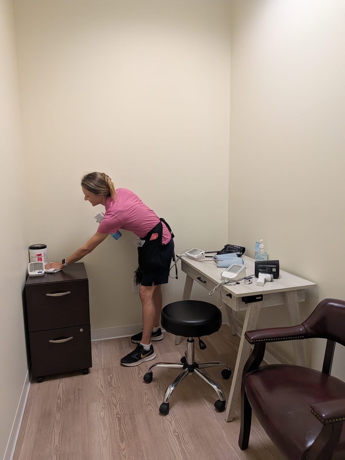 A cleaner in a pink polo wiping a file cabinet in a small room with a desk and blood-pressure monitors.