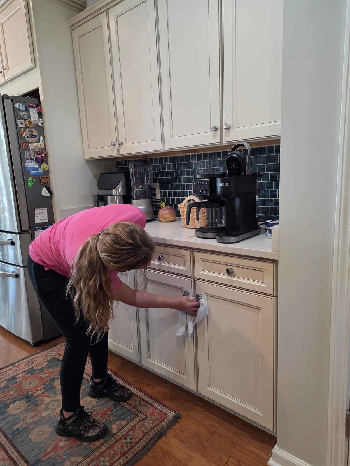 A cleaner in a pink polo wiping the lower kitchen cabinet doors.
