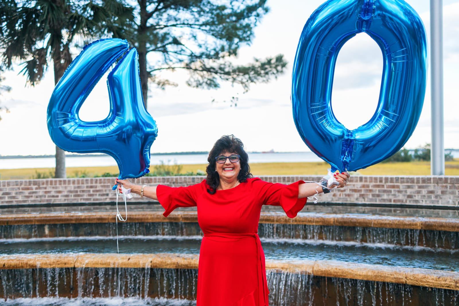 Teresa, the owner, in a red dress holding blue "4" and "0" balloons beside a fountain.