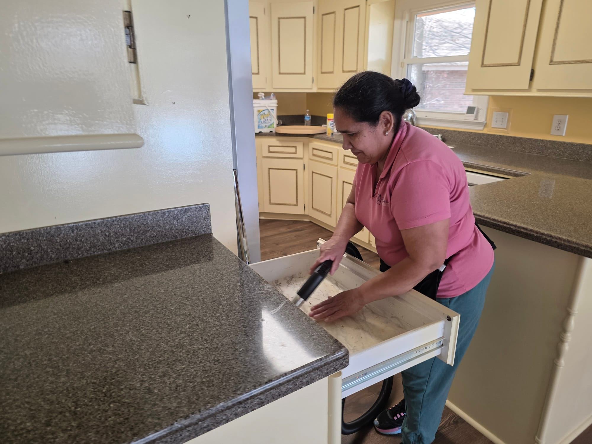 A cleaner in a pink polo vacuuming inside an open kitchen drawer.