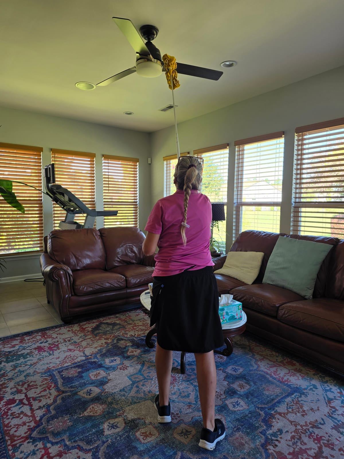 A cleaner in a pink polo dusting a ceiling fan with a long-handled duster in a sunroom.