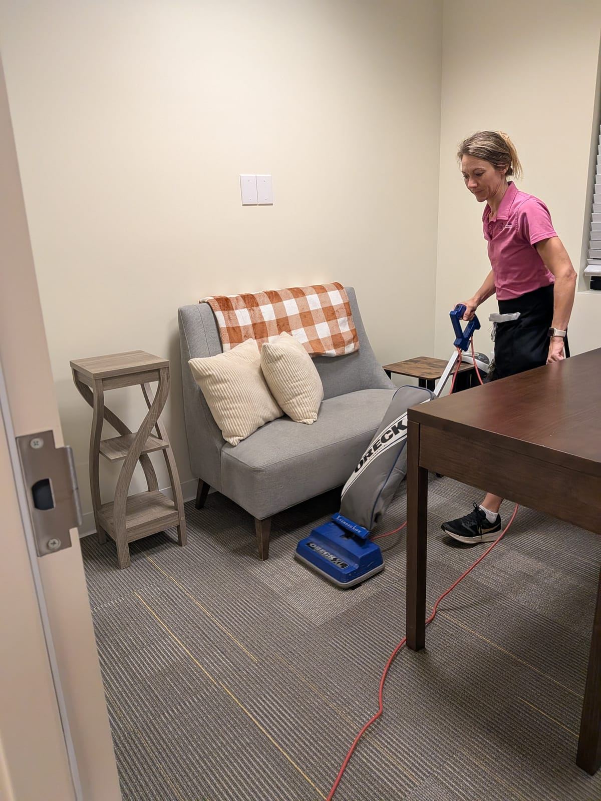 A cleaner in a pink polo vacuuming an office carpet with an upright vacuum.