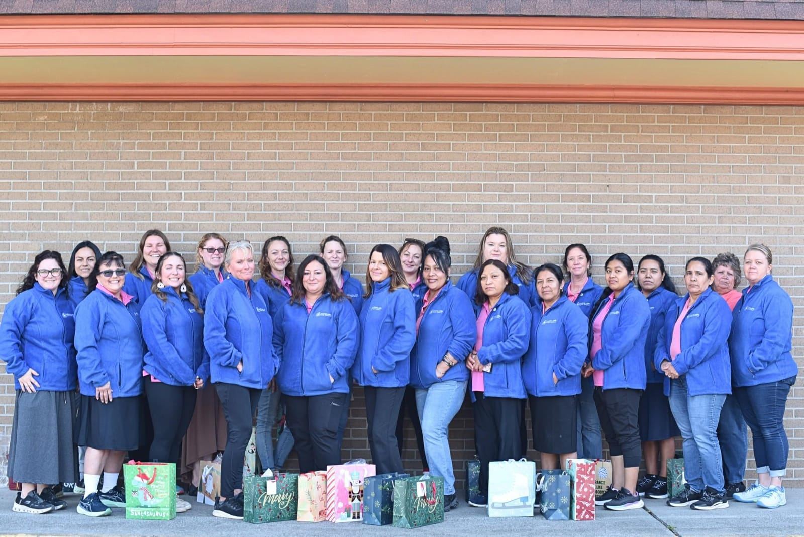 A large group of staff in blue fleece jackets standing in front of a brick wall with gift bags at their feet.