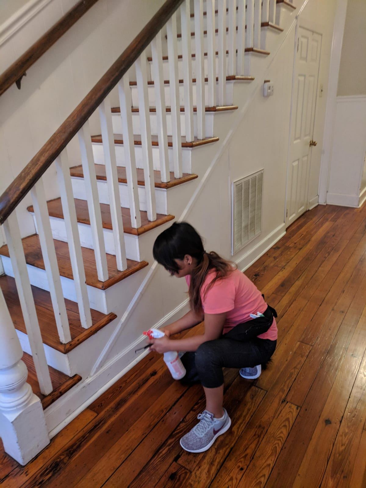 A cleaner in a pink shirt crouching to spray and wipe the side of a wooden staircase.