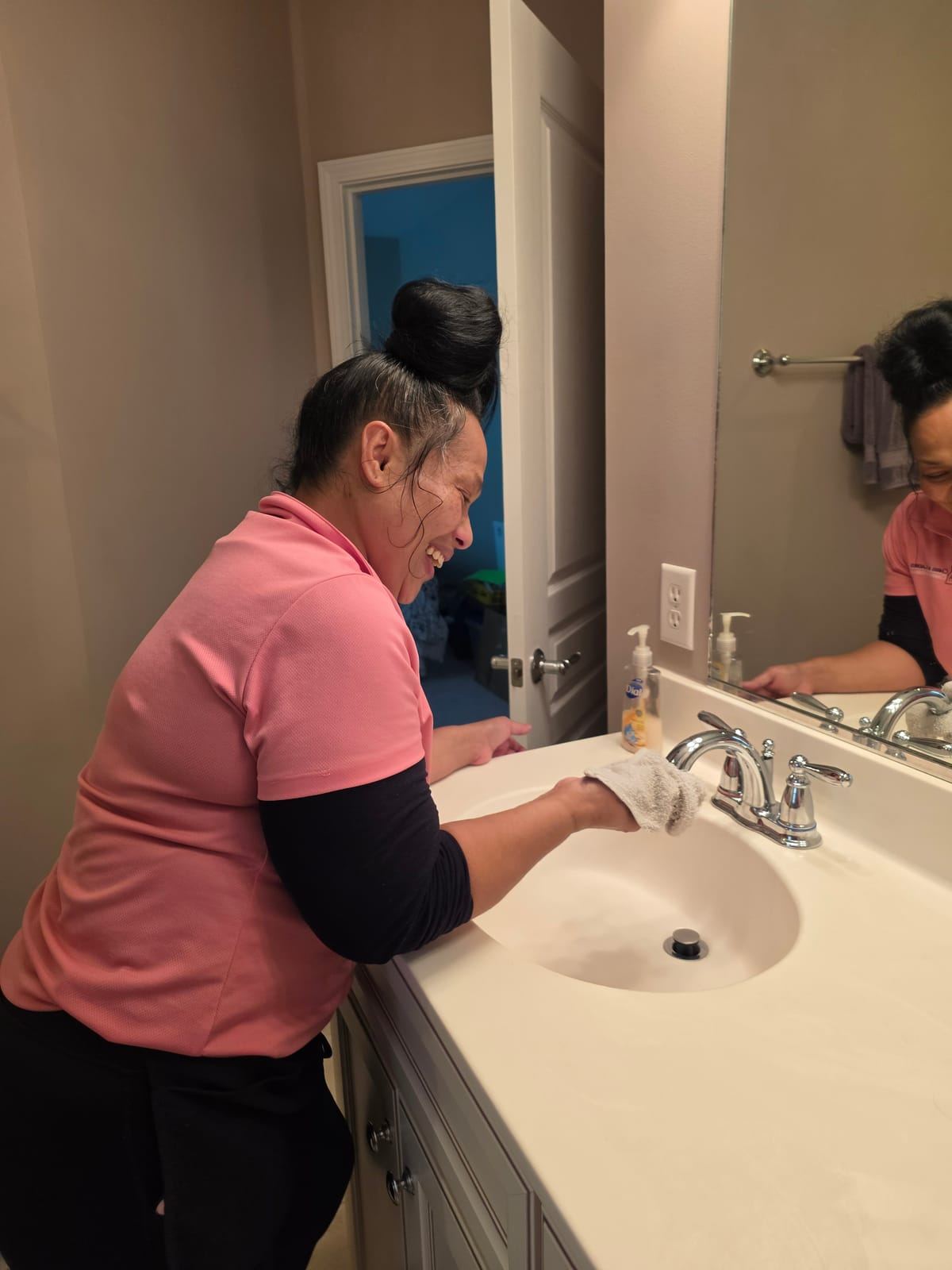 A smiling cleaner in a pink polo wiping a bathroom sink and faucet.