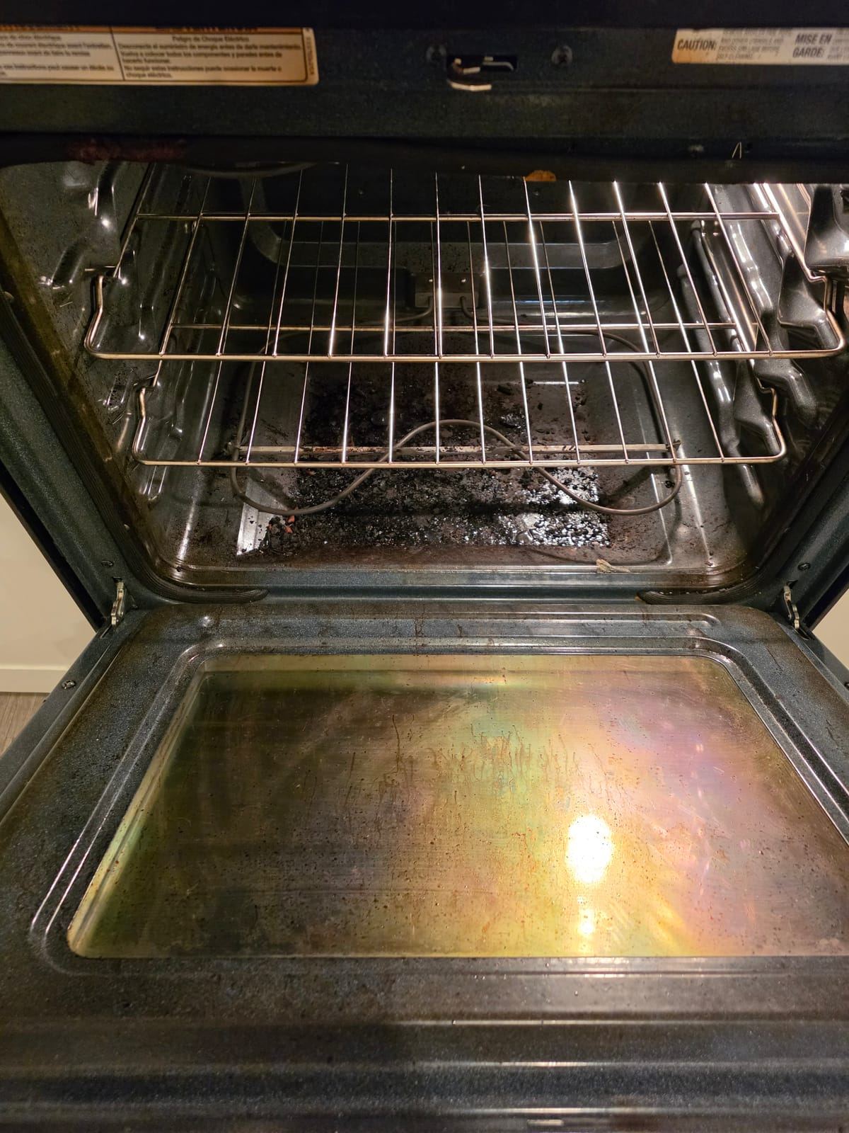 The inside of an open oven with burnt debris on the floor and a stained door glass.
