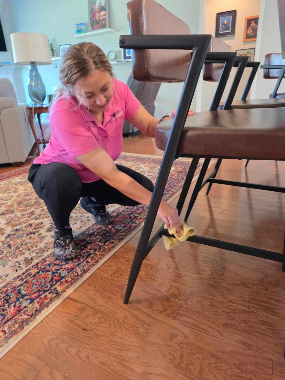 A smiling cleaner in a pink polo crouching to wipe the legs of a bar stool.