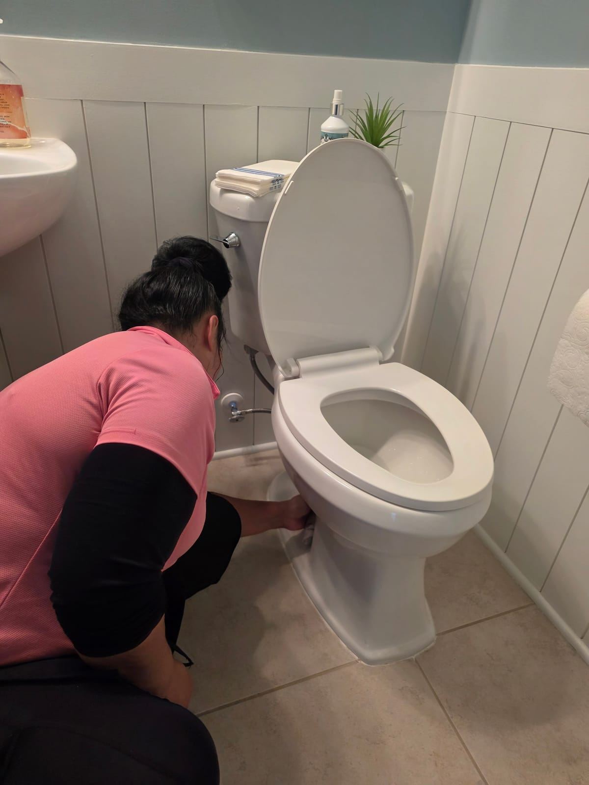 A cleaner in a pink polo crouching to wipe the base of a toilet.