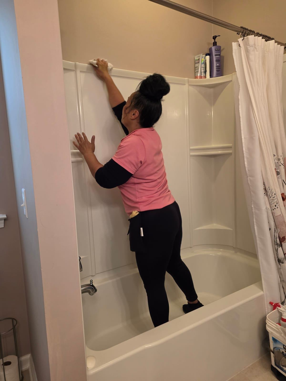 A cleaner in a pink polo standing in a bathtub wiping the top of the shower surround.