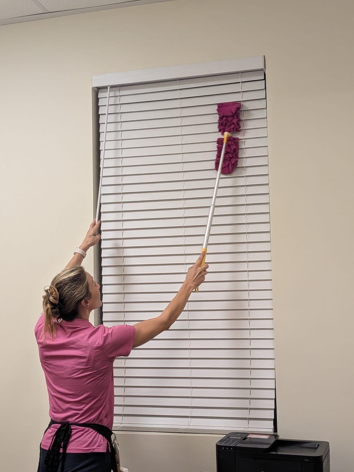 A cleaner in a pink polo dusting office window blinds with an extended duster.