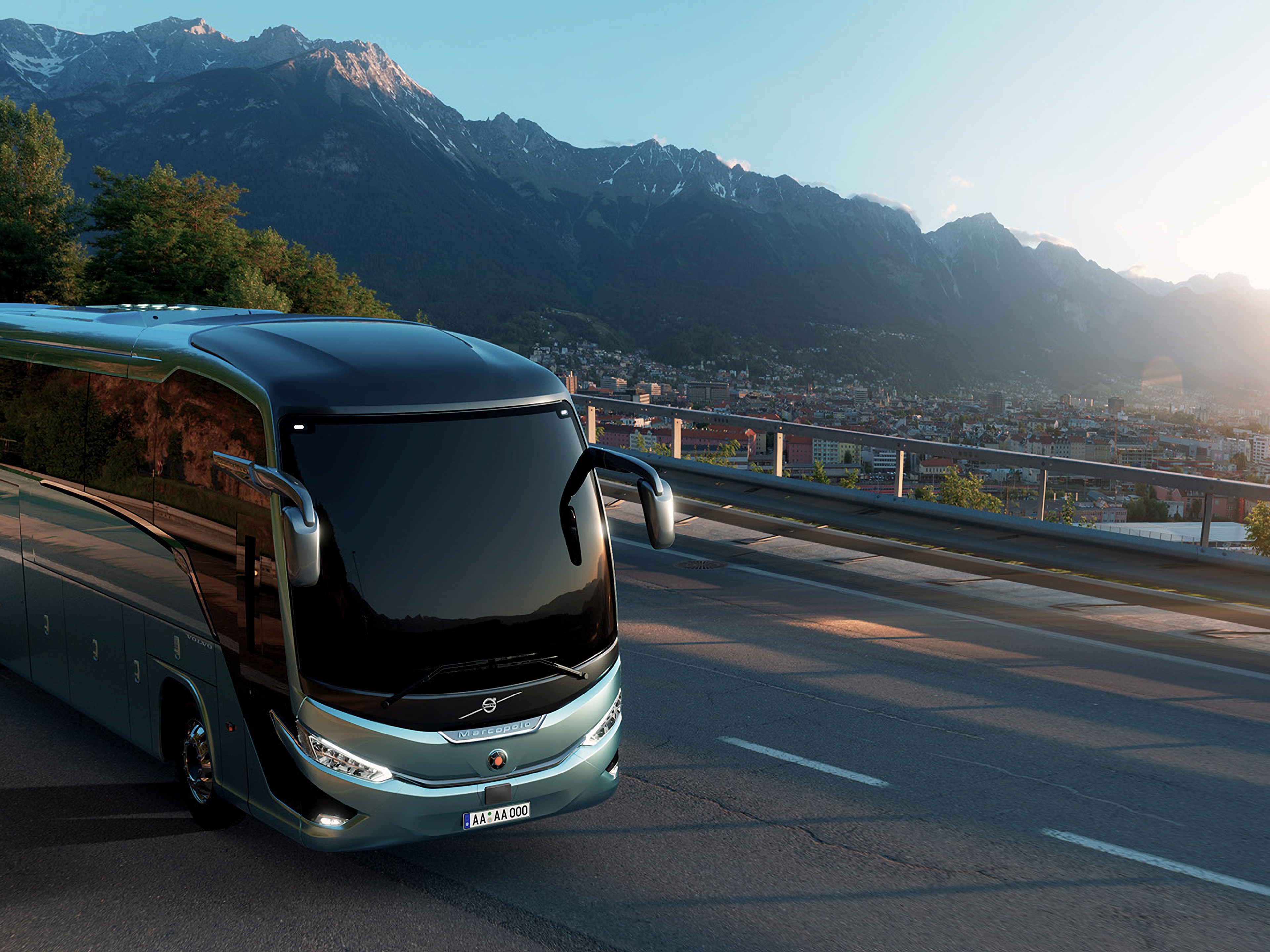 Modern bus driving along a mountain road overlooking a city at sunset.