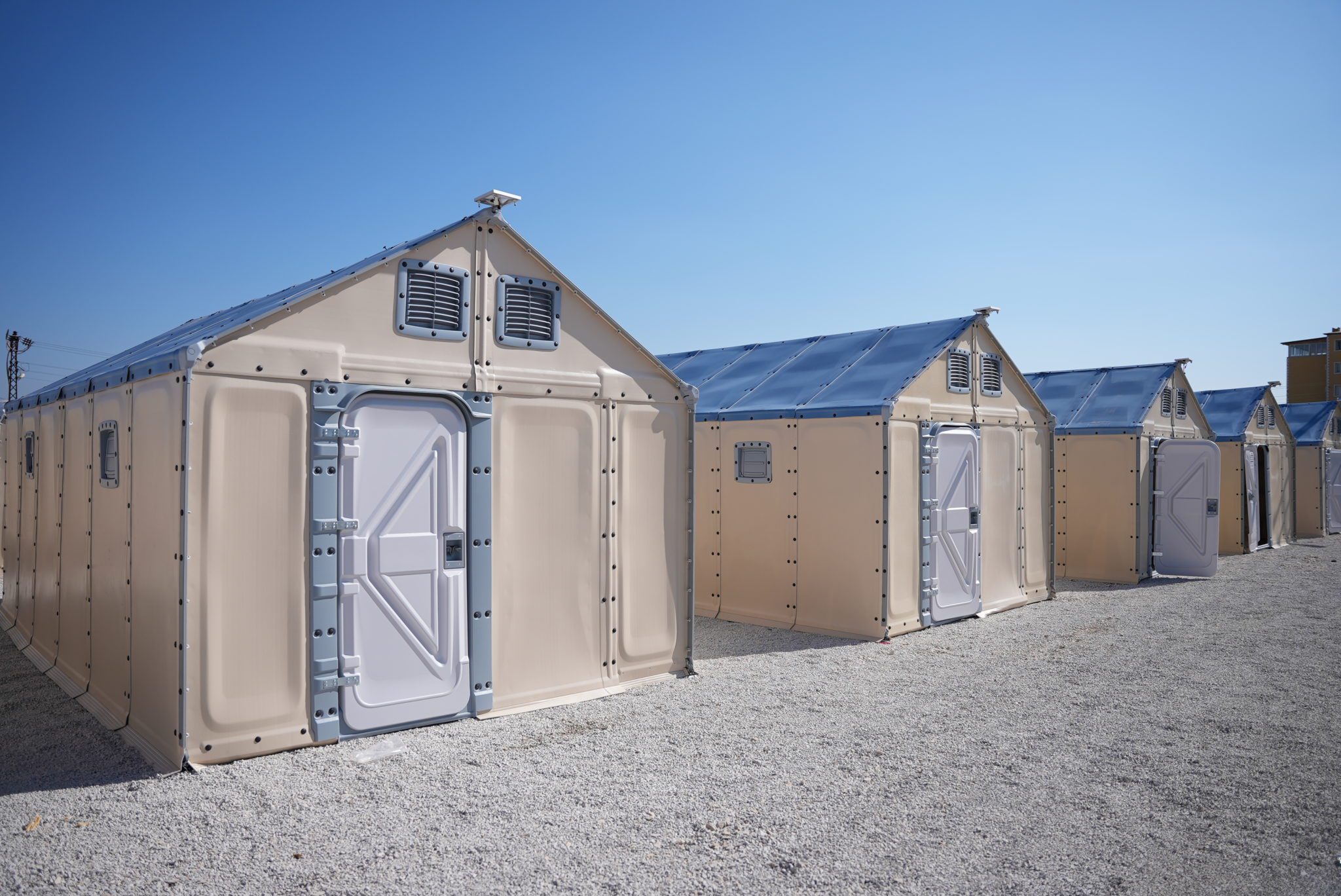 Row of modular shelter units with beige walls and white doors, set on gravel under a clear blue sky.