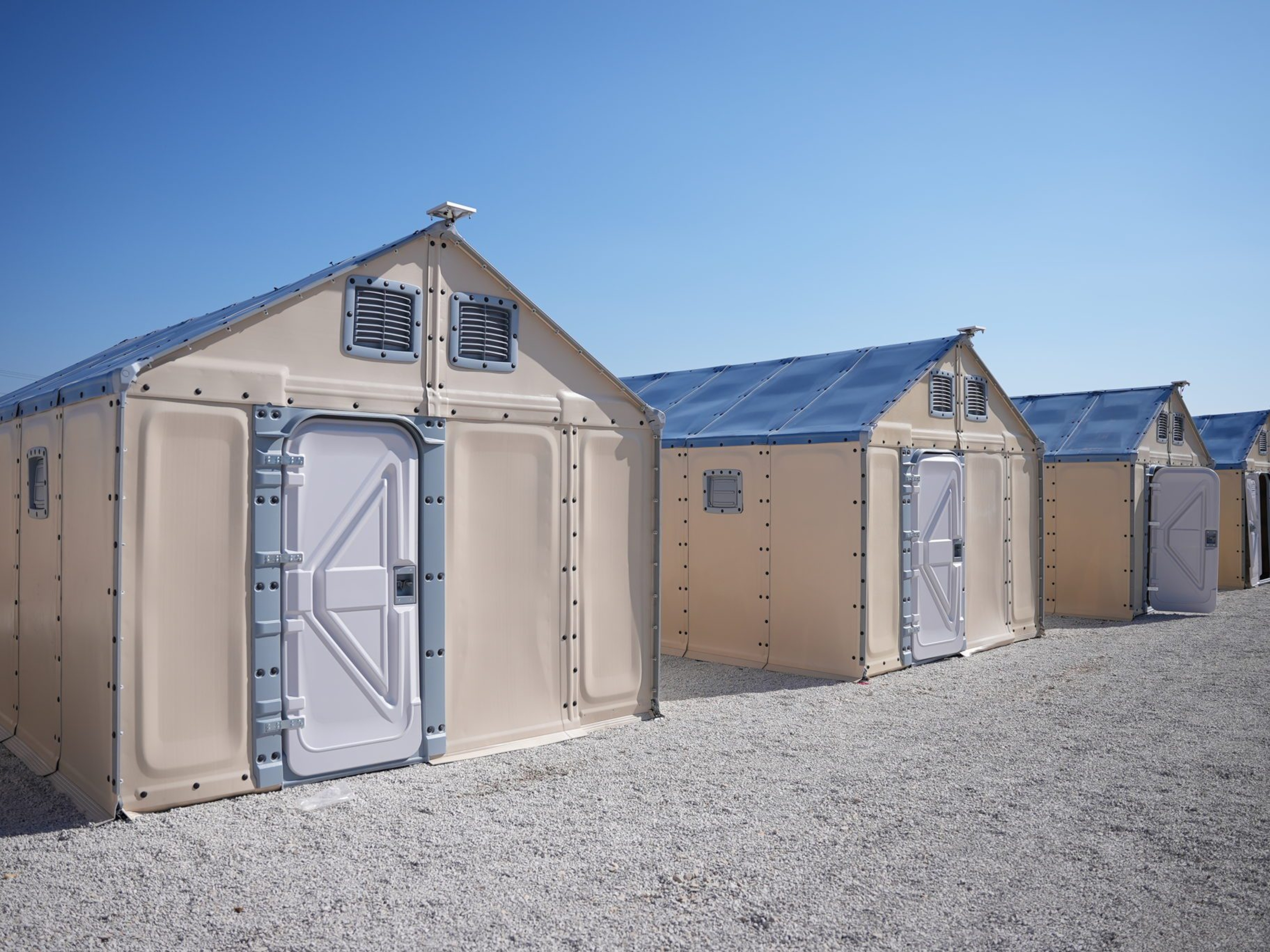 Row of modular shelter units with beige walls and white doors, set on gravel under a clear blue sky.