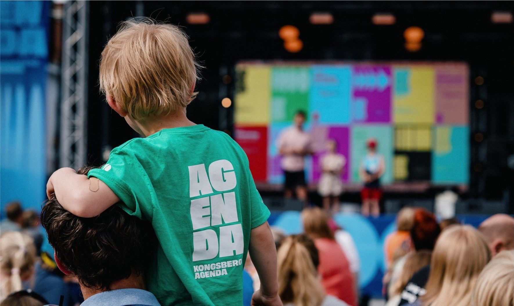 A boy sitting on a man shoulder and looking at a stage.