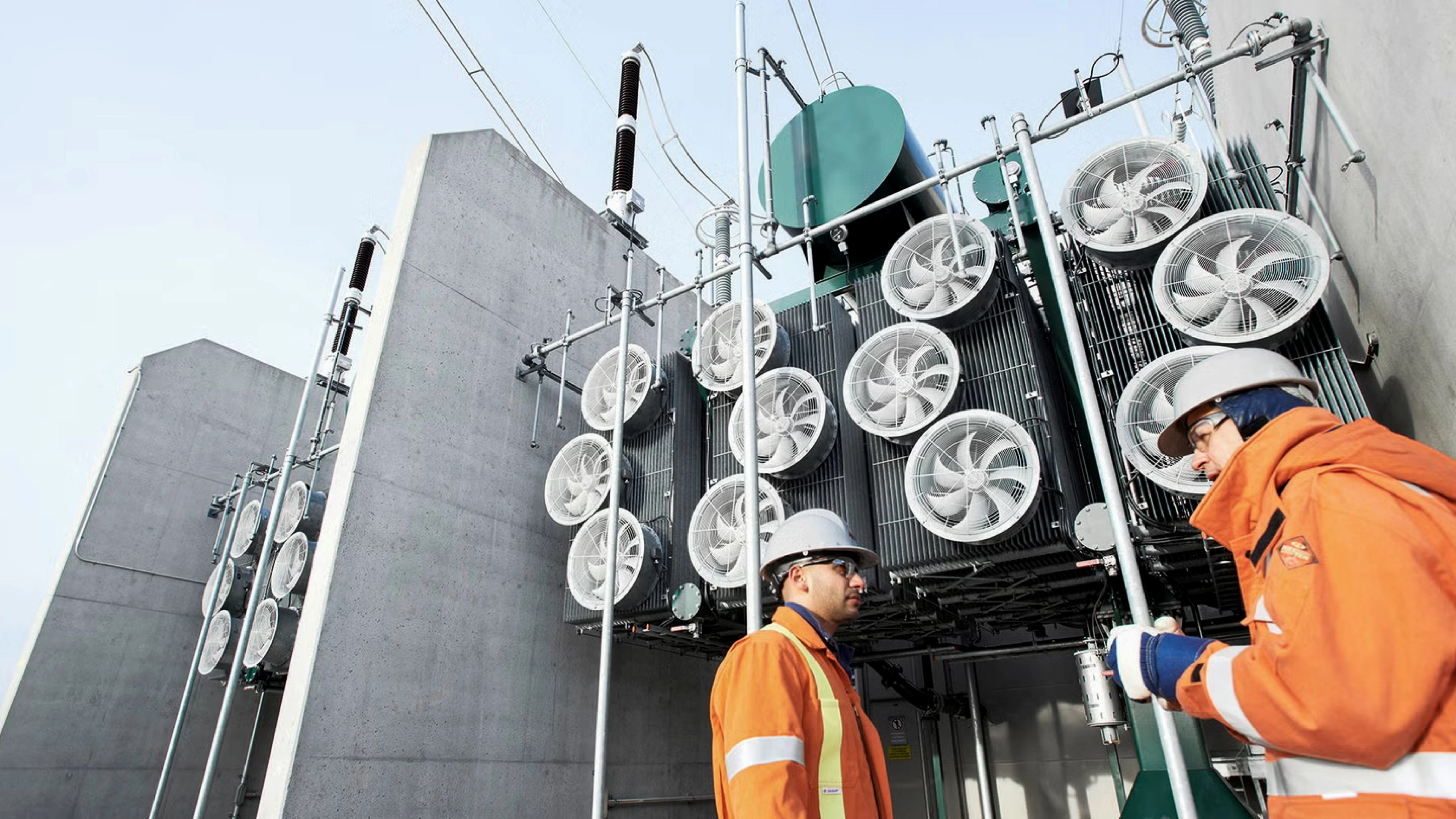Two technicians in orange high-visibility jackets and hard hats stand beside large industrial electrical equipment at an outdoor facility.
