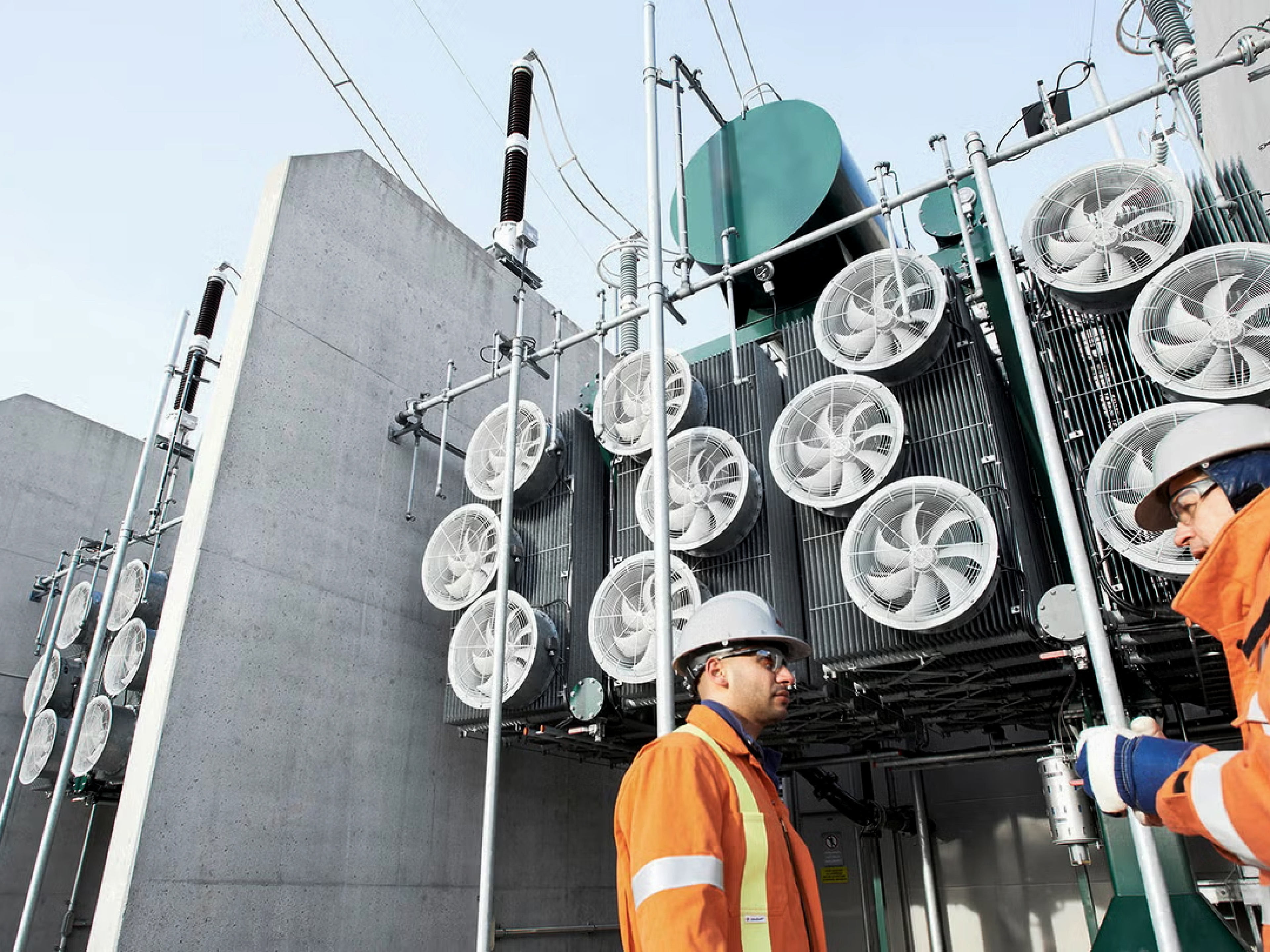 Two technicians in orange high-visibility jackets and hard hats stand beside large industrial electrical equipment at an outdoor facility.