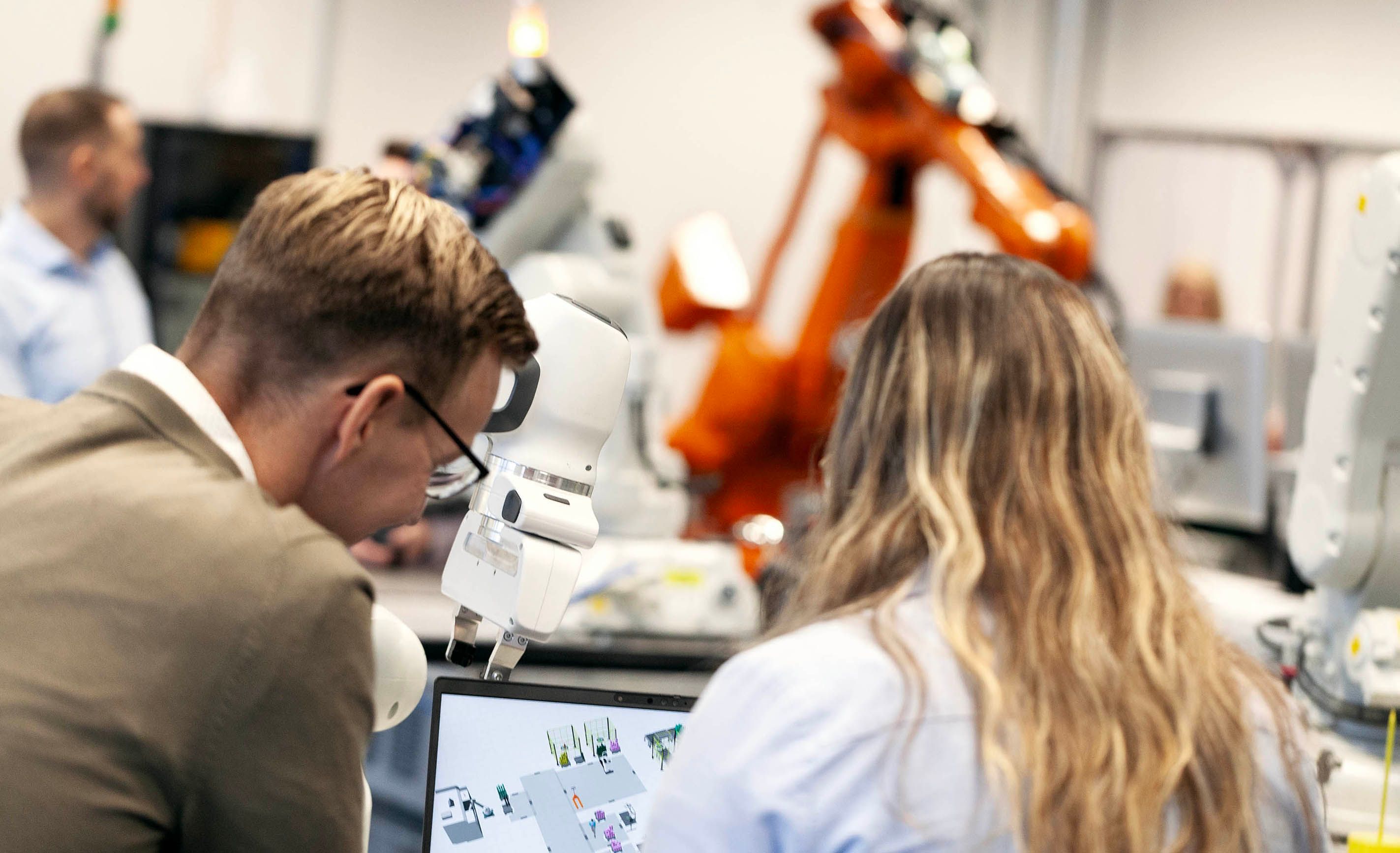 Two engineers working together at a workstation in an industrial lab with robotic arms in the background
