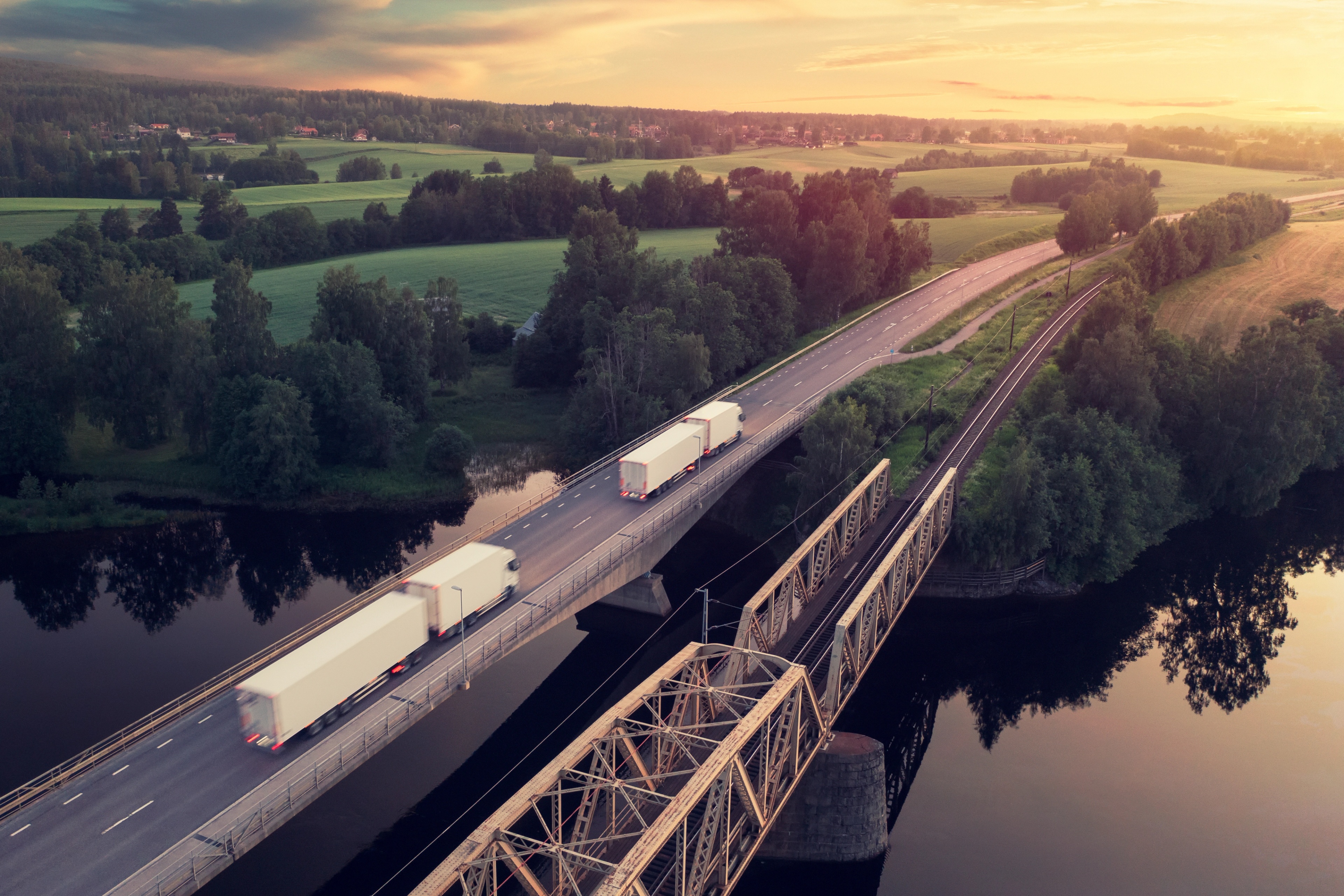 Trucks driving across a bridge over a river in a rural landscape at sunset.
