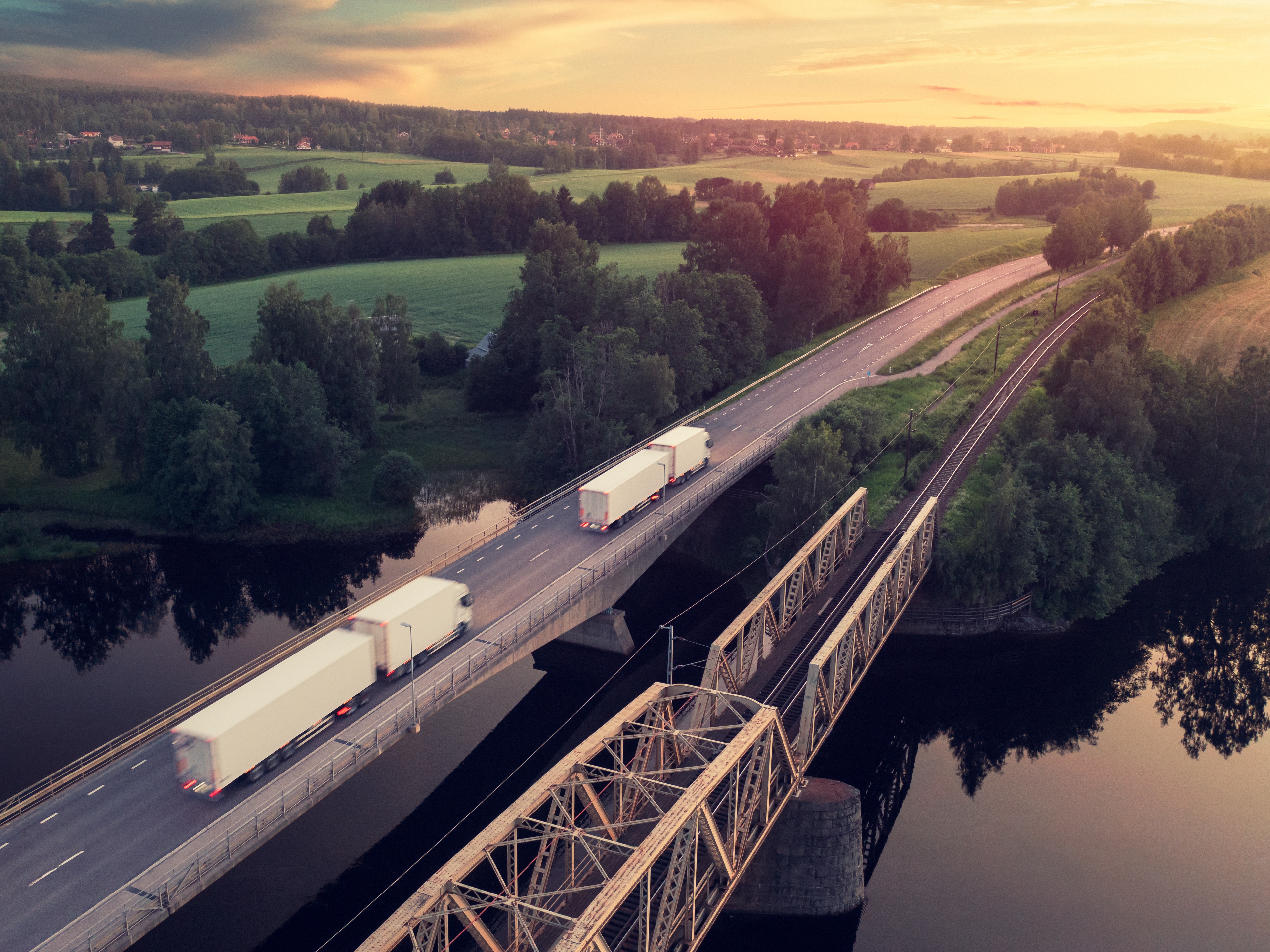 Trucks driving across a bridge over a river in a rural landscape at sunset.