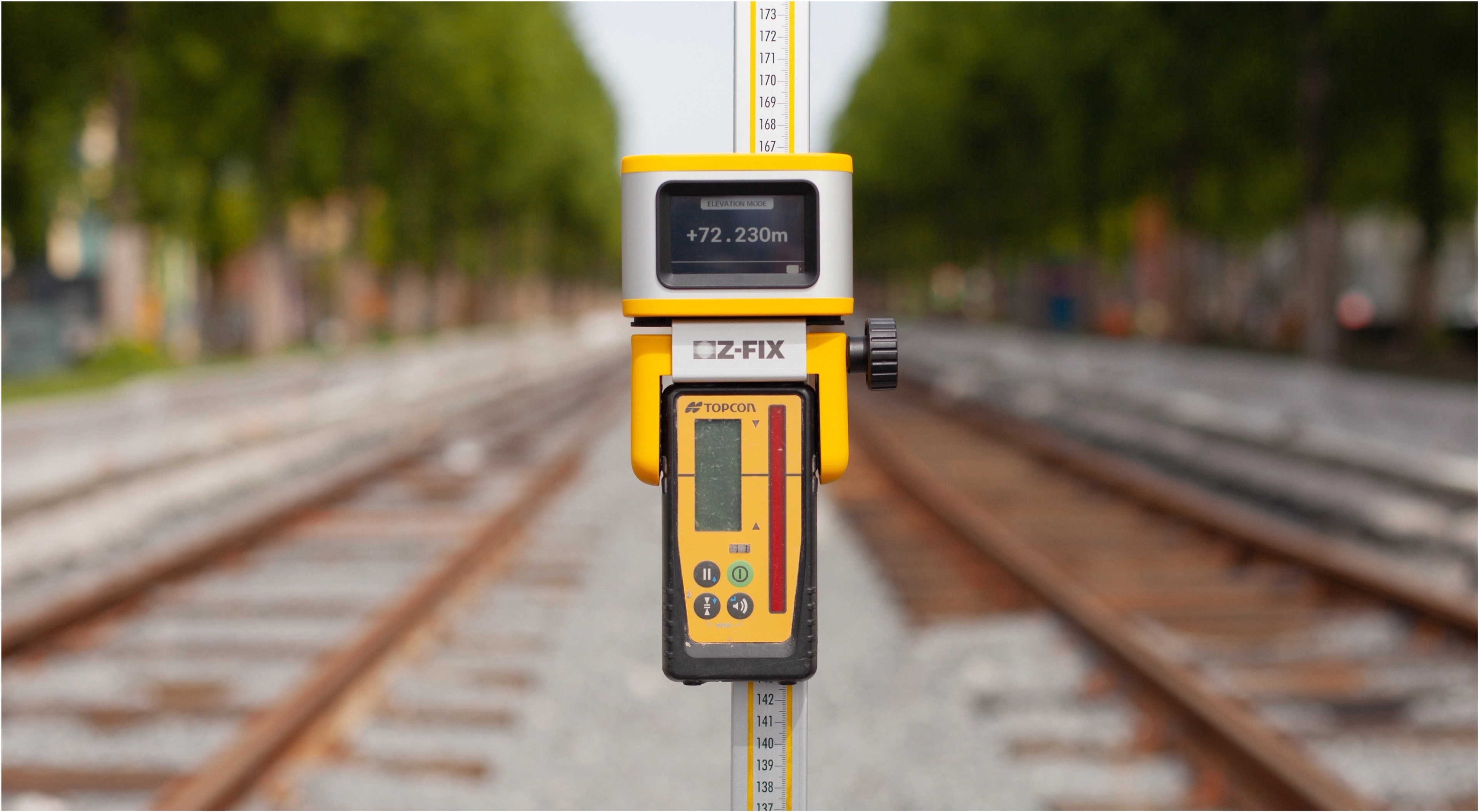 Close-up of a measuring tool mounted on a measuring rod, displaying elevation data, positioned on railway tracks with a blurred background.