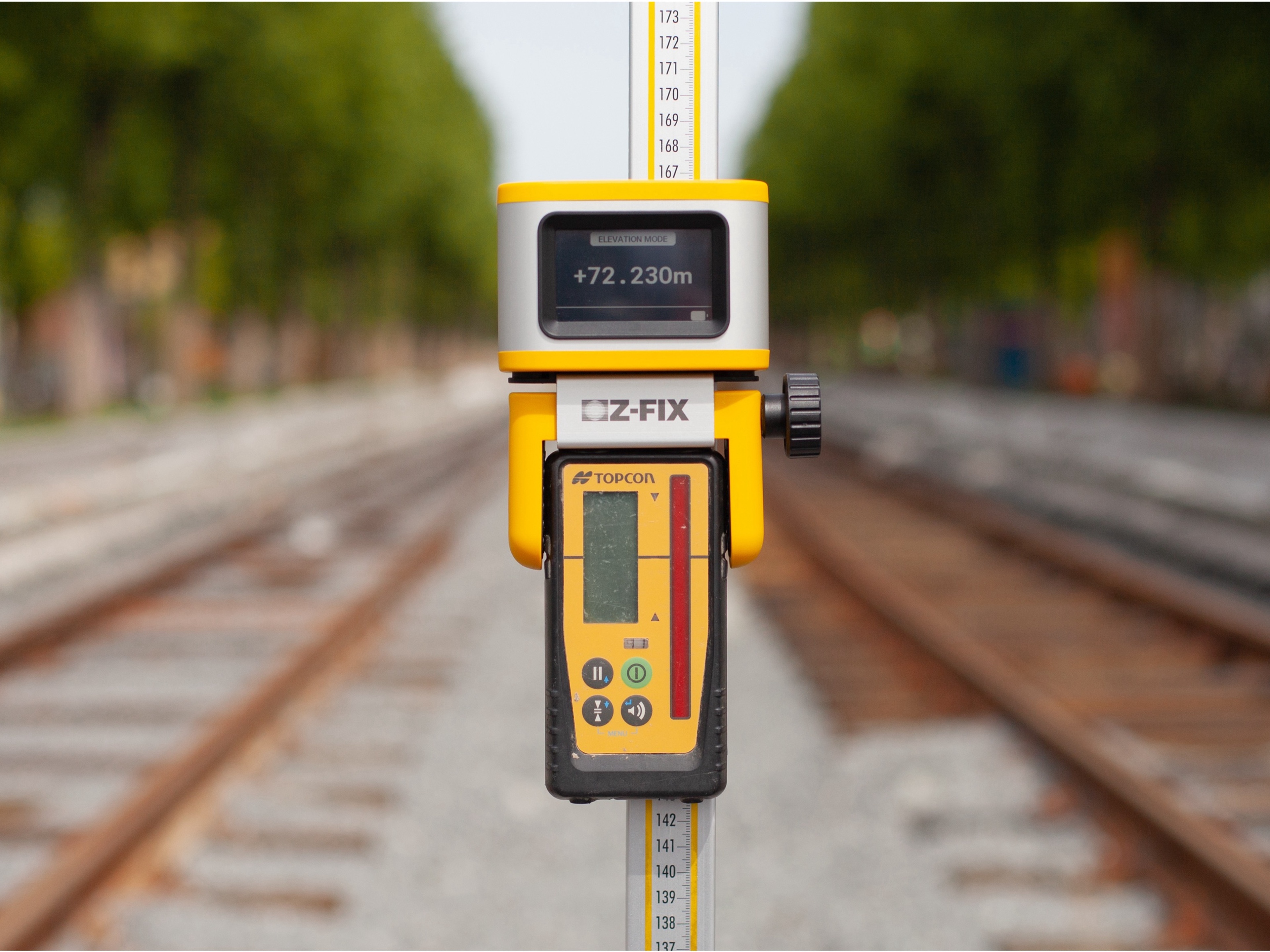 Close-up of a measuring tool mounted on a measuring rod, displaying elevation data, positioned on railway tracks with a blurred background.
