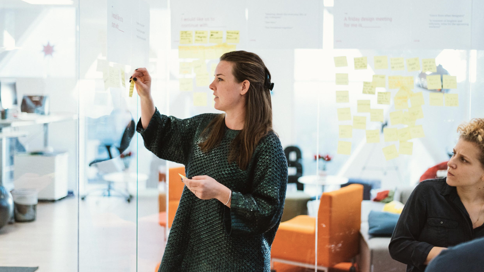 Two women in a room with glass walls, the walls are filled pith post-it notes