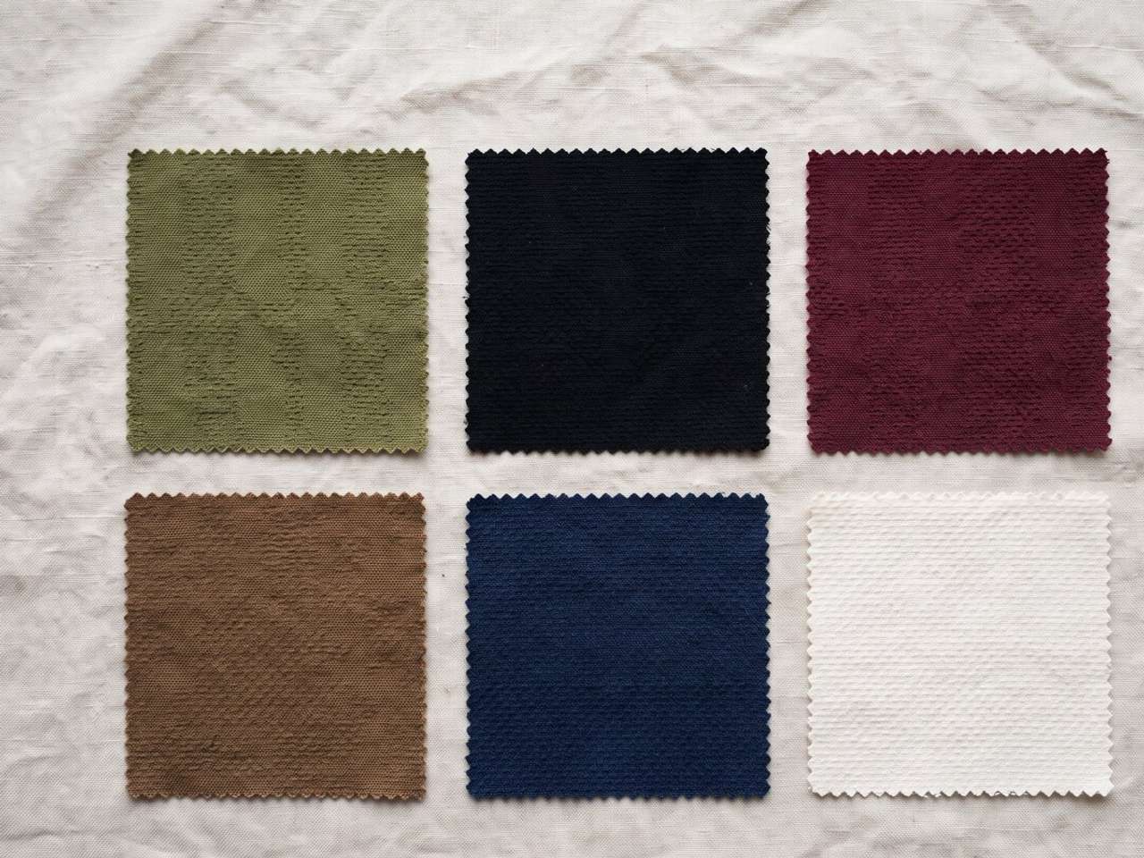 Six cotton dobby fabric swatches in olive, black, burgundy, brown, navy and off-white laid out on linen