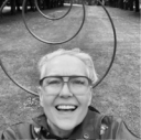 A black and white selfie image of a fair skinned woman with cropped hair and big glasses standing in front of an outdoor art installation.