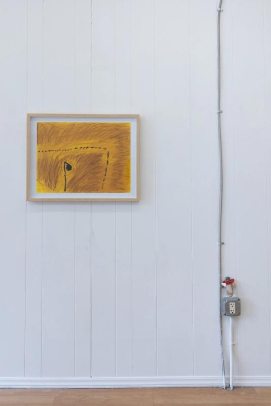 a view of two artworks. One is a framed drawing on yellow paper and focuses on a dark brown/black form that appears like a streetlight. going from left and turning down to the bottom middle is a dotted line that appears like a road or path. atop an electrical junction box is a small angel whose body is made of a mango pit. her head is a small gray stone and she wears a red ruffled collar.