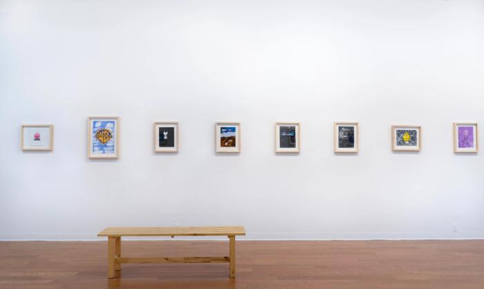 a white walled gallery with wooden floor and artworks in pale wooden frames on the wall in a single line. they are multicolored.