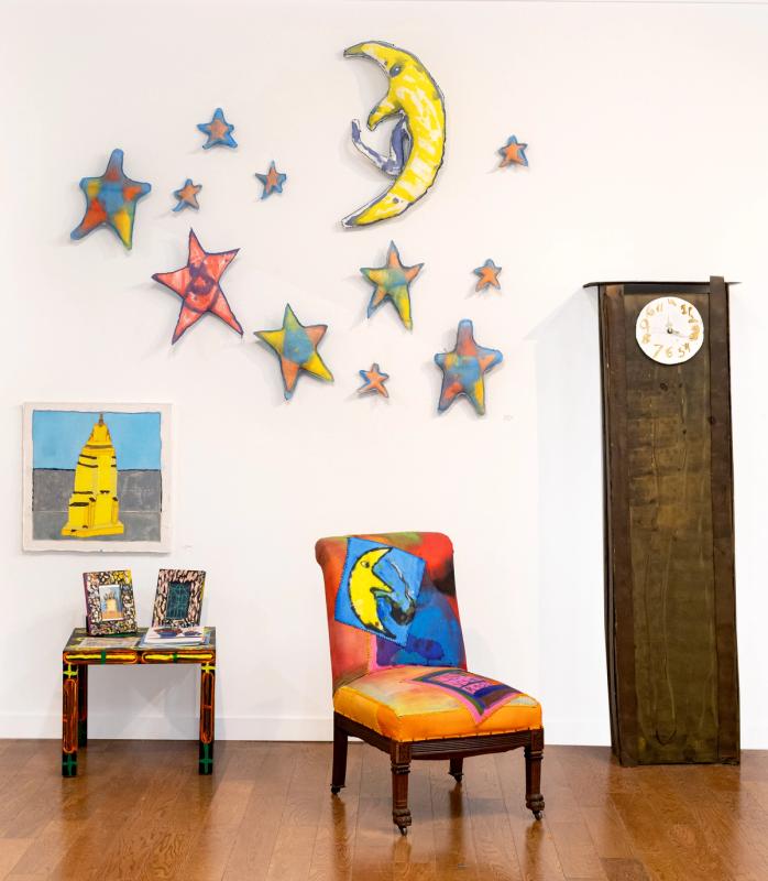 An install shot of the Living Room exhibition featuring a custom-made orange, pink, and red chair with an embroidered moon. A large, handmade cardboard sculpture resembling a grandfather clock and a series of moon and star-themed pillows hanging on the wall.