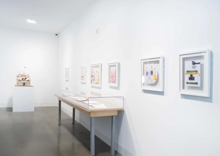 a view of a gallery space with white walls and grey floor. artworks are hung on the walls and there is a grey sculpture on a pedestal in the background, too small to make out detail but a house shape