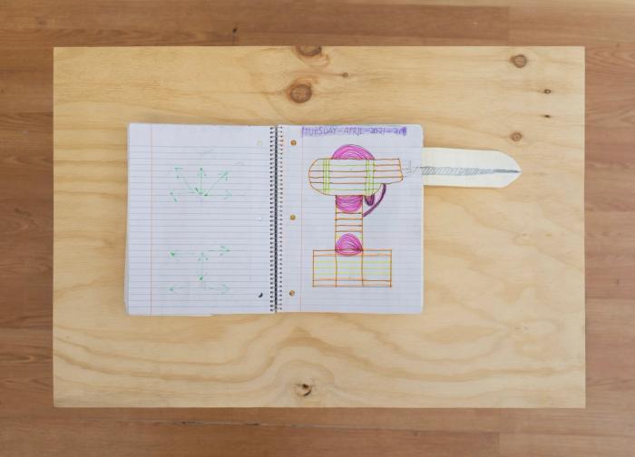 an overhead shot of an open spiral bound notebook. the page we can see has an addition in a long thin shape. on the page is a drawing of a drill in bright pink, yellow, and orange highlighter. the silver drill tip goes off the page and continues along the addition thats been taped on.